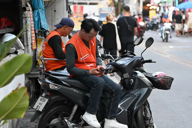 DFC 3742 Two delivery riders in orange vests sit on and beside a parked scooter along a busy street one checking his phone while passersby and traffic move in the background